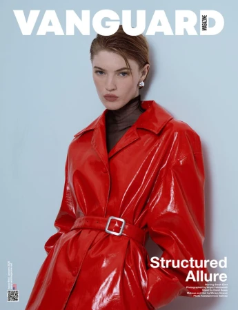 Model in a glossy red belted coat against a plain pale background for Vanguard Magazine