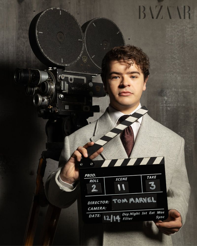 Adult male poses with a vintage film camera and a clapperboard in a photo studio; he wears a suit in front of a grey wall.