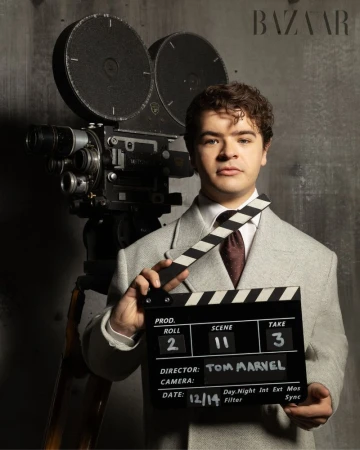 Adult male poses with a vintage film camera and a clapperboard in a photo studio; he wears a suit in front of a grey wall.