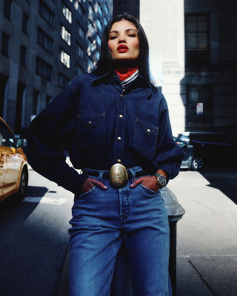 A woman poses in denim shirt and jeans with red lipstick and a patterned scarf against an urban background with a yellow taxi.