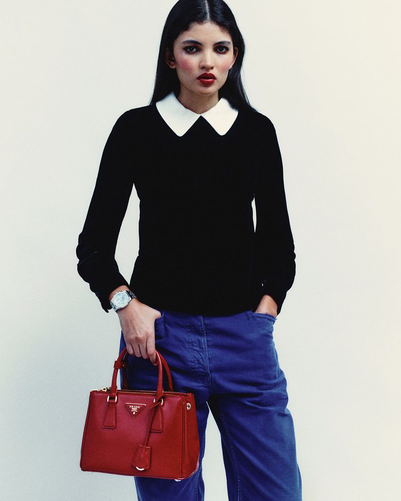 Woman in black sweater and blue pants holds a red leather Prada handbag against a white background in a fashion editorial shot.