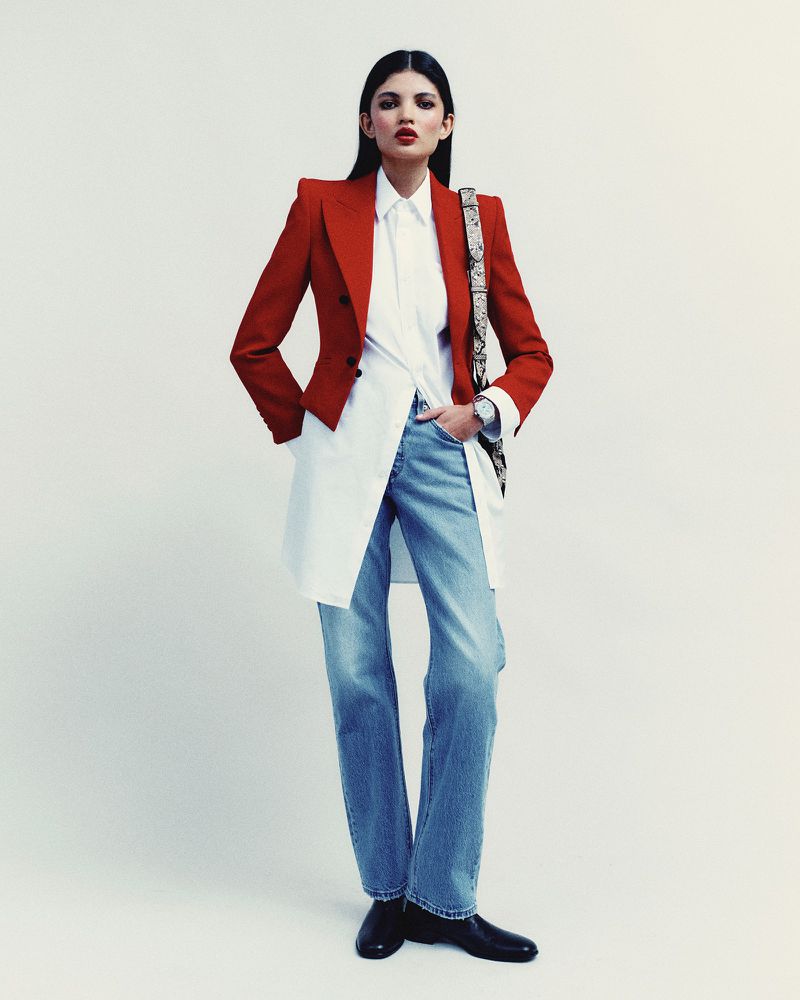 A woman with long dark hair stands against a white background, wearing a red tailored blazer over a long white shirt and light blue jeans.