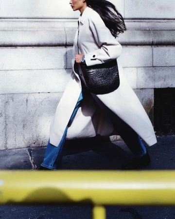 Model in long white coat and blue jeans walking past a stone wall for Vogue Latin America