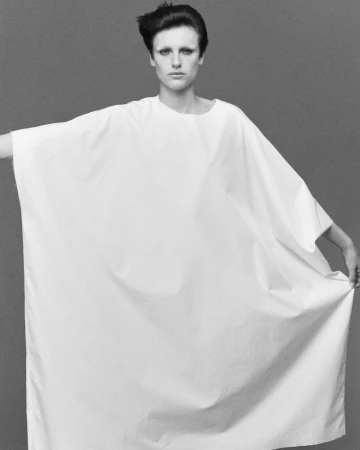 Black and white portrait of a model in a simple, draped white dress, posing against a neutral gray background in a fashion studio.