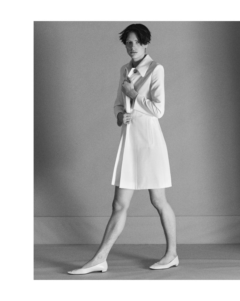 Monochrome full shot of androgynous fashion model in coat dress and flats walking against a grey backdrop.