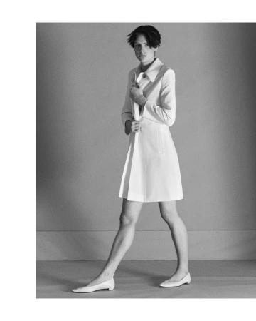 Monochrome full shot of androgynous fashion model in coat dress and flats walking against a grey backdrop.