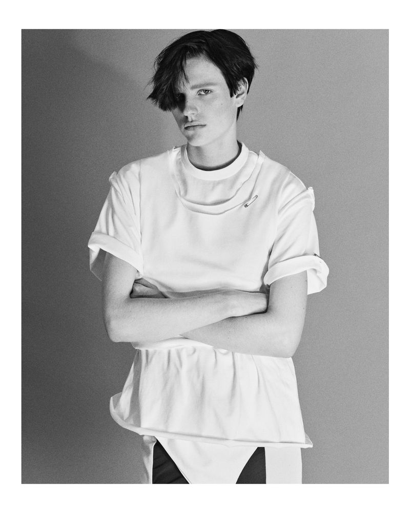 Black and white portrait of an adult female with dark, short hair in a white t-shirt with her arms crossed in front of a grey background.