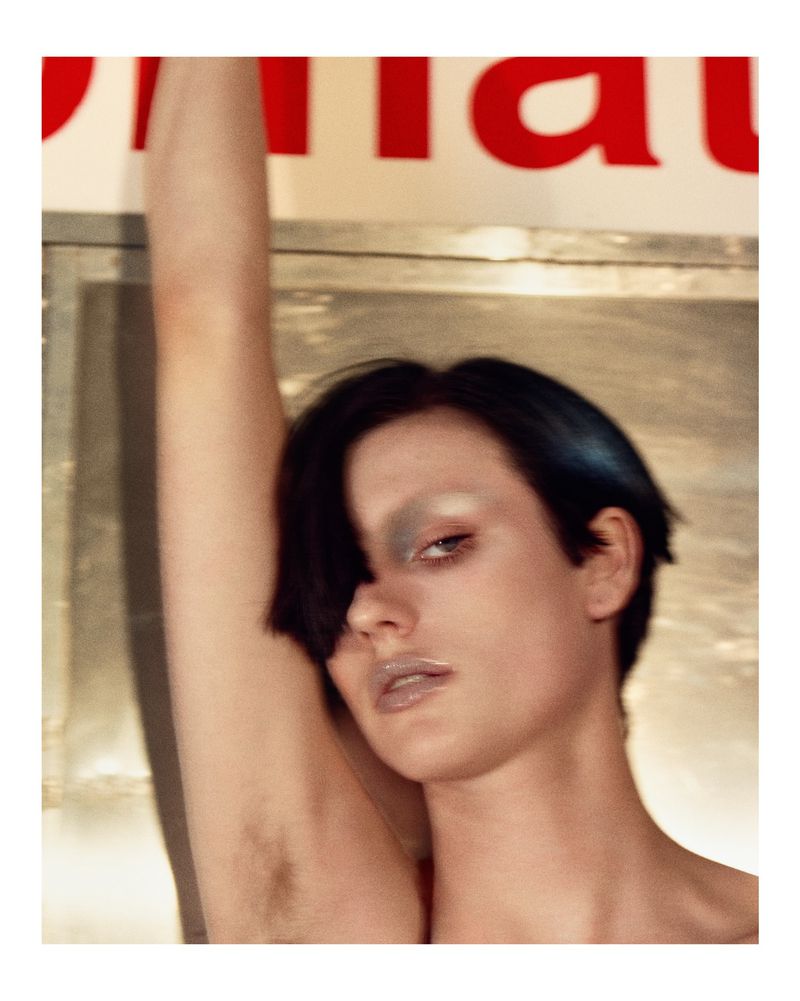 Close-up studio shot of a fair-skinned adult female model with an underarm pose, bold silver eyeshadow, glossy lips, and short dark hair.