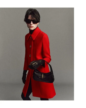 A female model with sunglasses poses in a red coat and black gloves, carrying a black leather purse against a gray background.