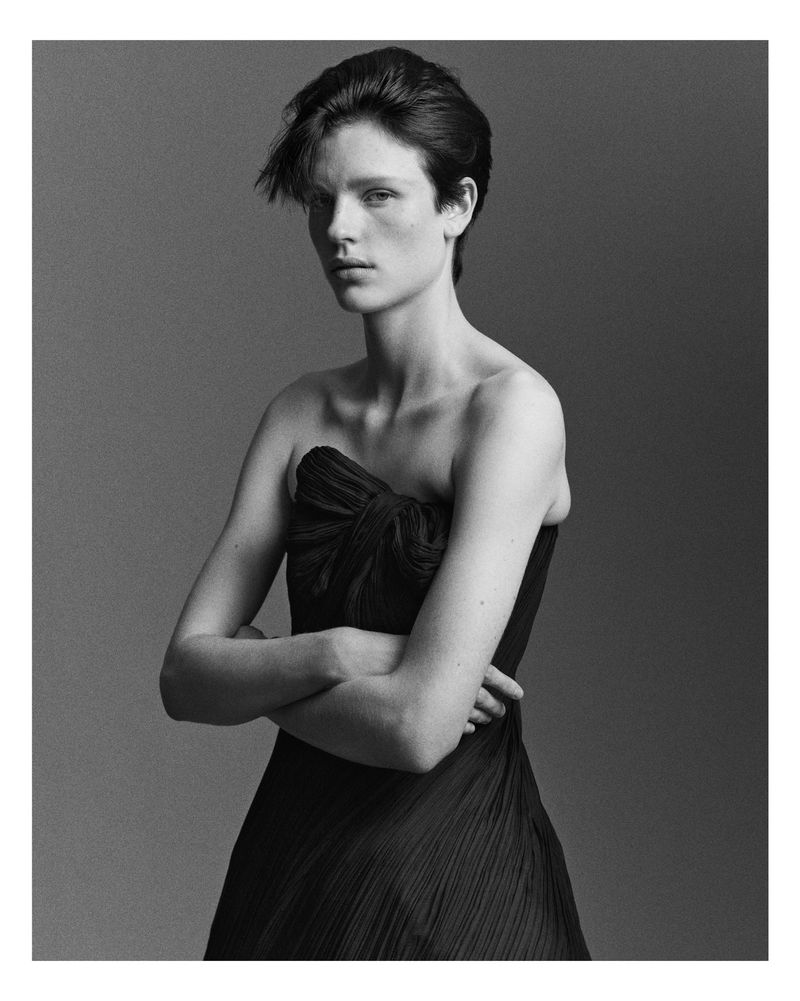 Black and white portrait of a young adult woman in a draped strapless dress, posing in a photo studio with arms crossed.