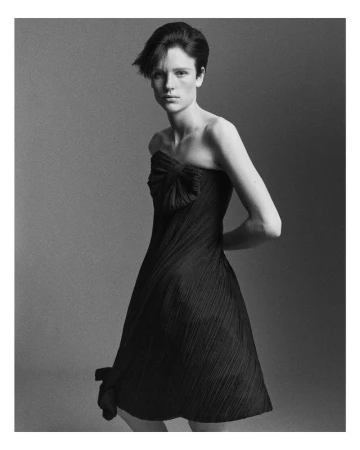 A single woman in a black strapless dress poses against a gray backdrop, hands behind her back. Black-and-white fashion studio shot.