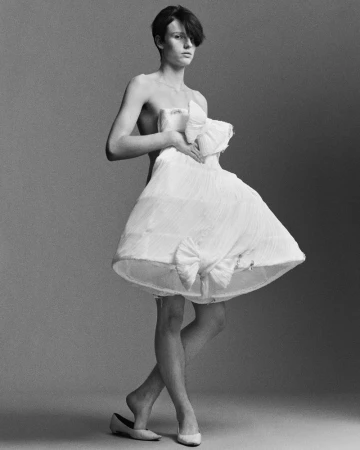 A person stands in a photo studio, wearing a strapless dress and ballet shoes, holding the dress away from their body.