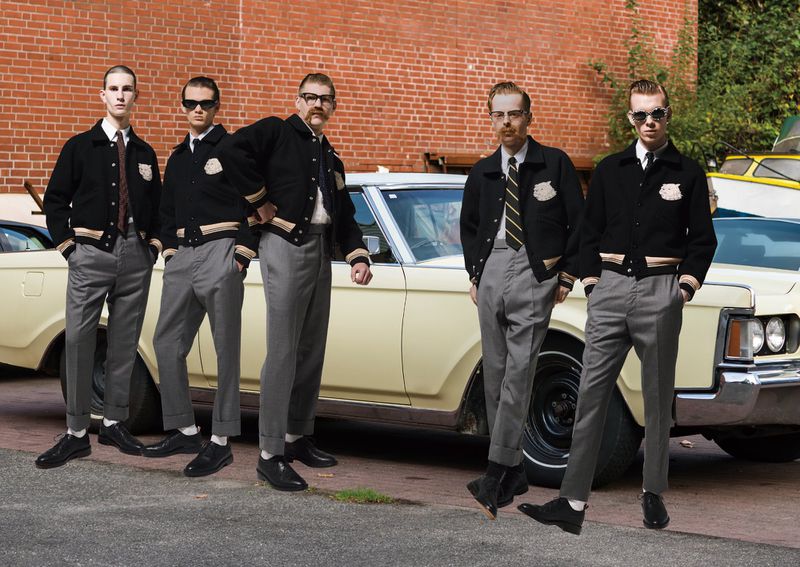 Five young adult men stand in front of an old yellow car, wearing matching black jackets and grey pants, against a brick wall backdrop.