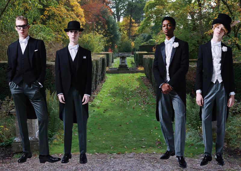 Four men in formal morning suits pose in a verdant outdoor garden with trimmed hedges and trees providing a classic and elegant style.