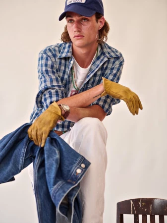 A young adult male poses indoors wearing a blue plaid shirt, white pants, and a blue baseball cap, holding a denim jacket and gloves.