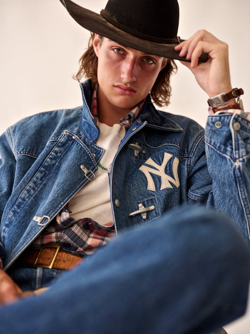 Portrait of a young adult man wearing a dark hat and denim jacket with an embroidered logo, seated indoors with a light background.