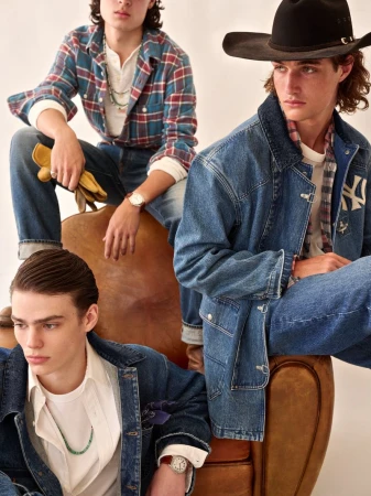 Three young adult men in Western-inspired denim fashion sit on and around a vintage leather chair in a studio setting.