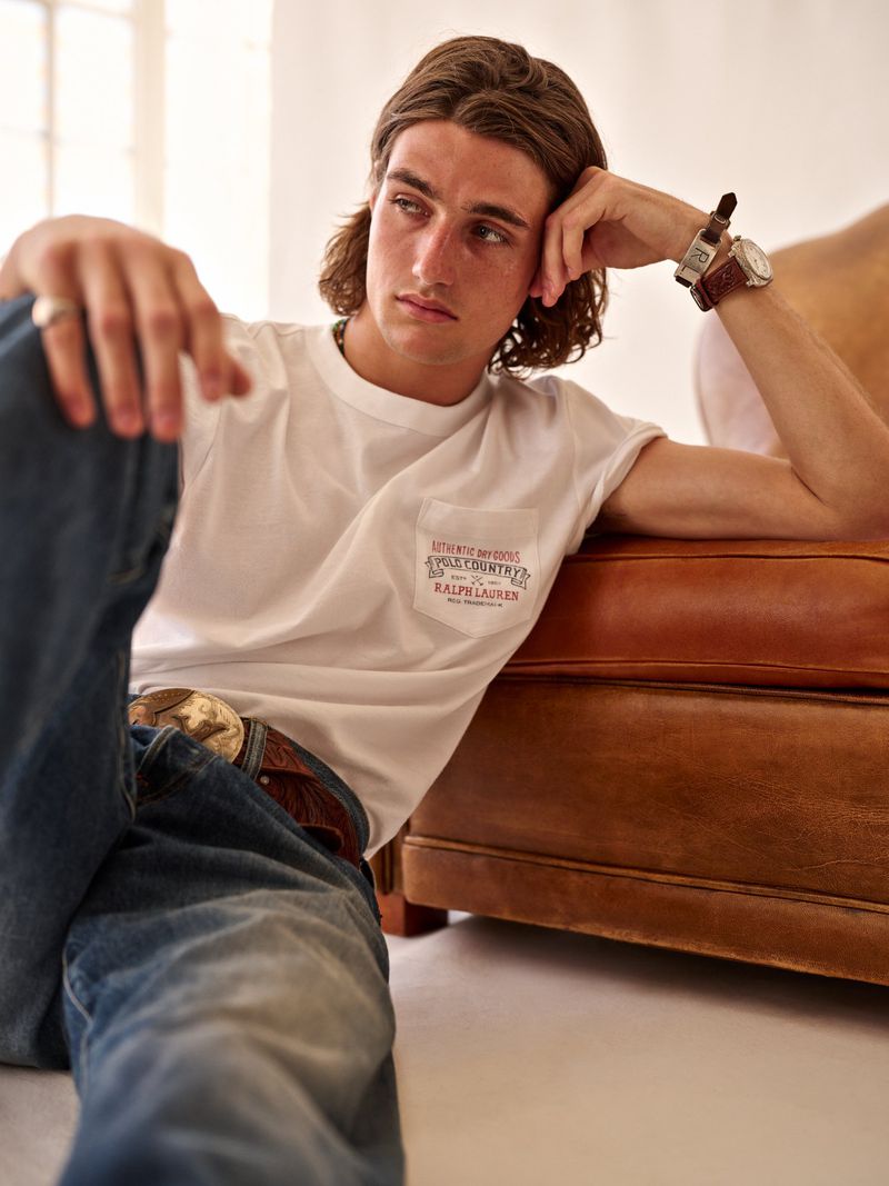 Man in a white t-shirt leans against a brown leather couch in a bright interior. He's wearing jeans and a watch. Casual fashion.