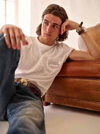 Man in a white t-shirt leans against a brown leather couch in a bright interior. He's wearing jeans and a watch. Casual fashion.