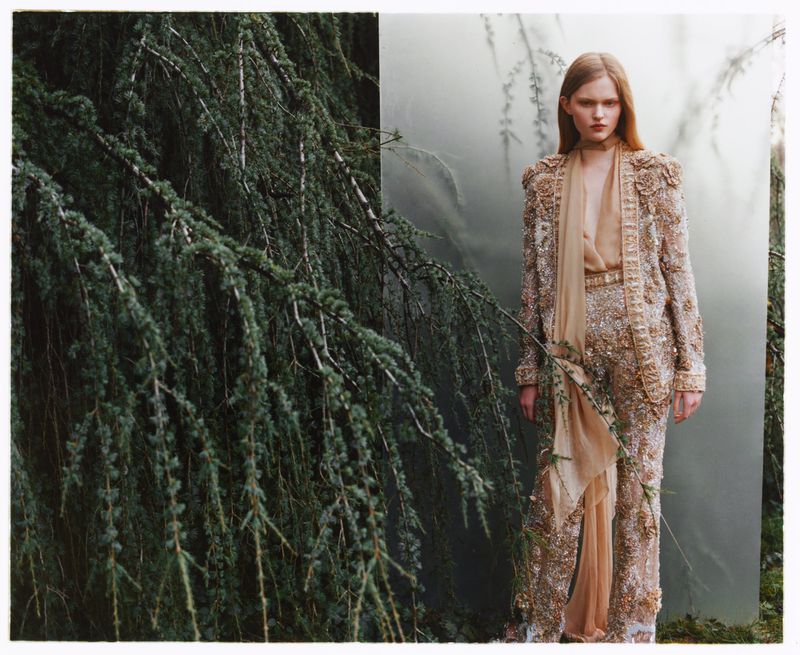 Woman in an embellished tan pantsuit stands in front of a forest and tree branches, in a fashion editorial style.