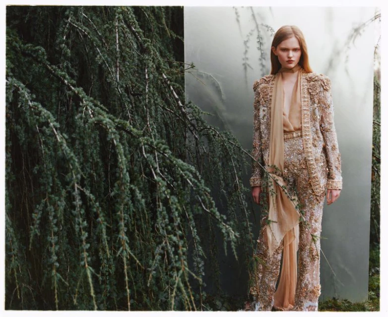 Woman in an embellished tan pantsuit stands in front of a forest and tree branches, in a fashion editorial style.