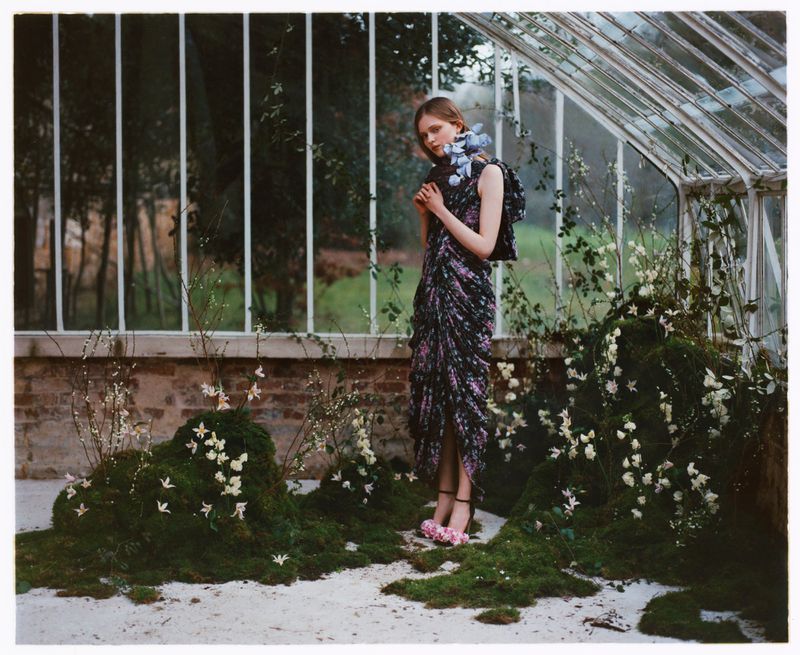 Fashion shot of a young woman in a floral dress with floral accents in a sunlit greenhouse, surrounded by moss and climbing vines.