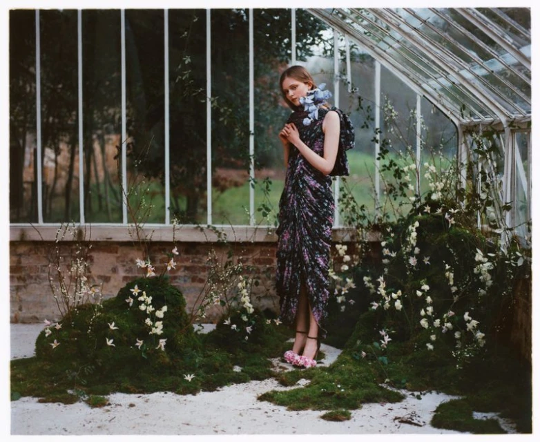 Fashion shot of a young woman in a floral dress with floral accents in a sunlit greenhouse, surrounded by moss and climbing vines.