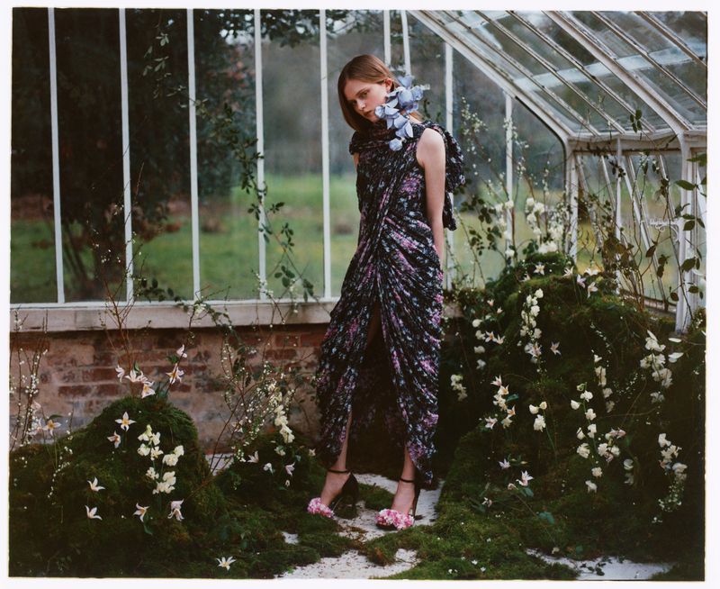 Elegant adult woman in floral dress posing near a greenhouse with moss and flower decorations, capturing an outdoor fashion editorial.