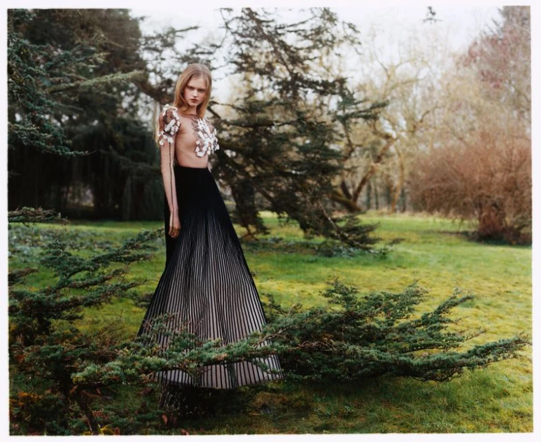 A fashionable woman stands in a grassy park, wearing a sheer top with floral embellishments and a long, pleated skirt, posing amongst trees.