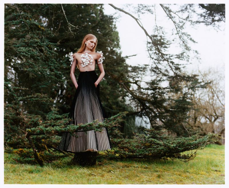 Fashion shot of a fair-skinned adult woman in a partly sheer floral top and black-to-grey striped skirt posing in a lush green outdoor setting.