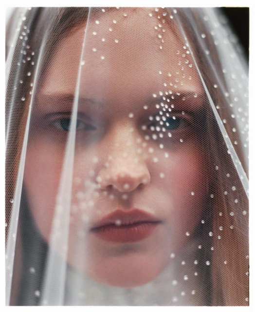 Close-up portrait of a young, fair-skinned adult female with brown hair, wearing a white beaded veil, looking directly at the camera.
