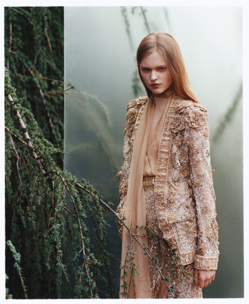 A fair-skinned woman with light brown hair poses in a lush outdoor setting. She wears a beaded tan suit with a sheer scarf.