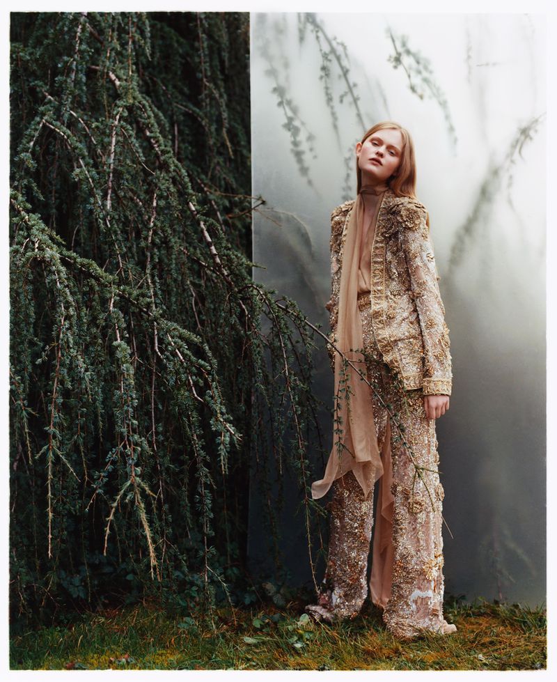 A woman poses in an ornate outfit, standing near a tree in a soft, dreamlike outdoor setting for a fashion editorial.