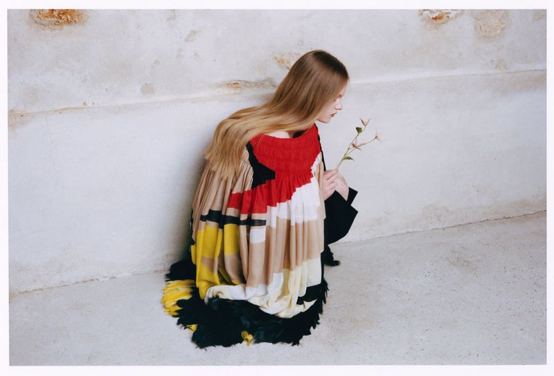 Model Ava Shipp in a colorful pleated haute couture garment holding a flower against a weathered plaster wall for Vogue China.