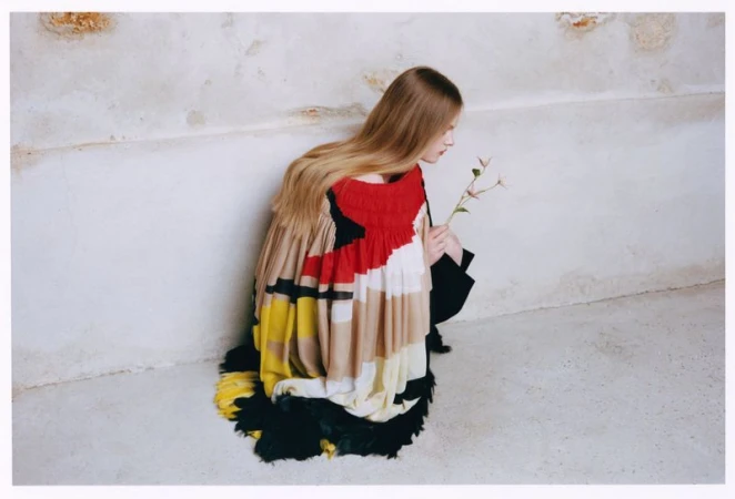 Model Ava Shipp in a colorful pleated haute couture garment holding a flower against a weathered plaster wall for Vogue China.