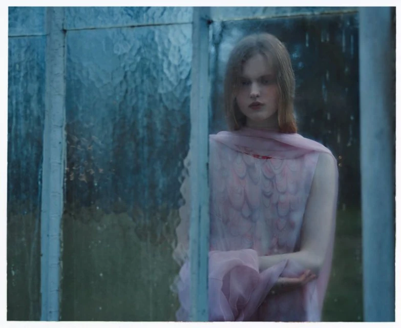 A young woman with fair hair and a pink dress stands behind a textured glass window in an indoor environment, creating an ethereal mood.