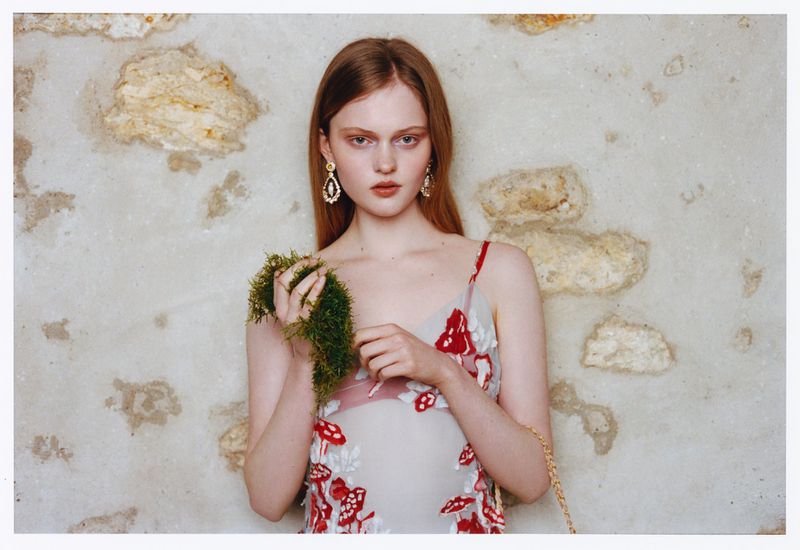 Elegant woman in sheer mushroom-embroidered dress poses in front of a textured stone wall wearing statement earrings and holding green moss.