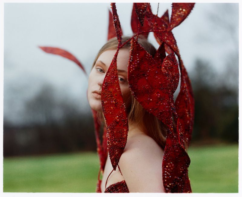 Portrait of a woman with red ornamental leaves framing her face in an outdoor setting, captured in a fashion editorial style.