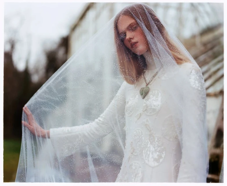 Close-up shot of an adult female model in an ornate white dress with a sheer veil, outdoors with muted natural lighting, fashion editorial.