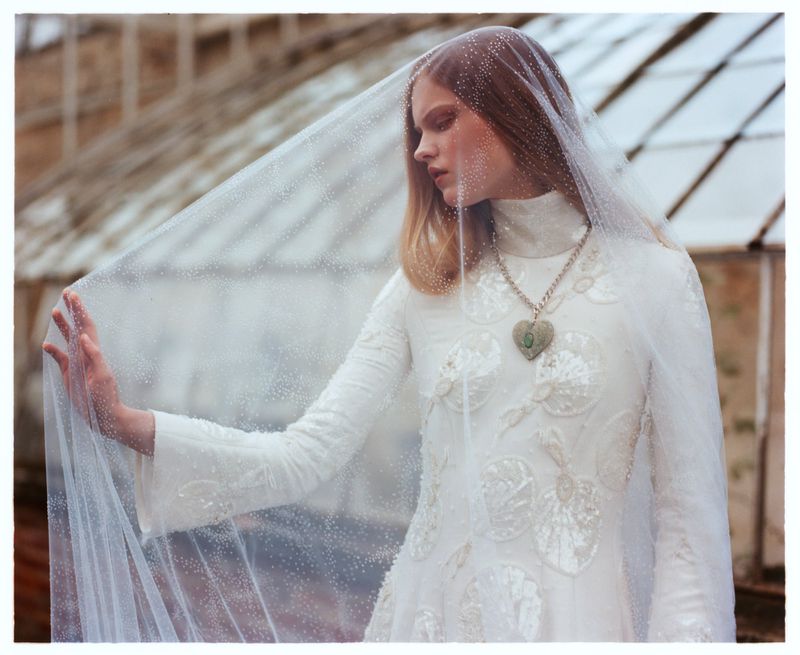 A fair-skinned woman with long blonde hair and a delicate white veil poses in a long-sleeved white dress against a greenhouse backdrop.