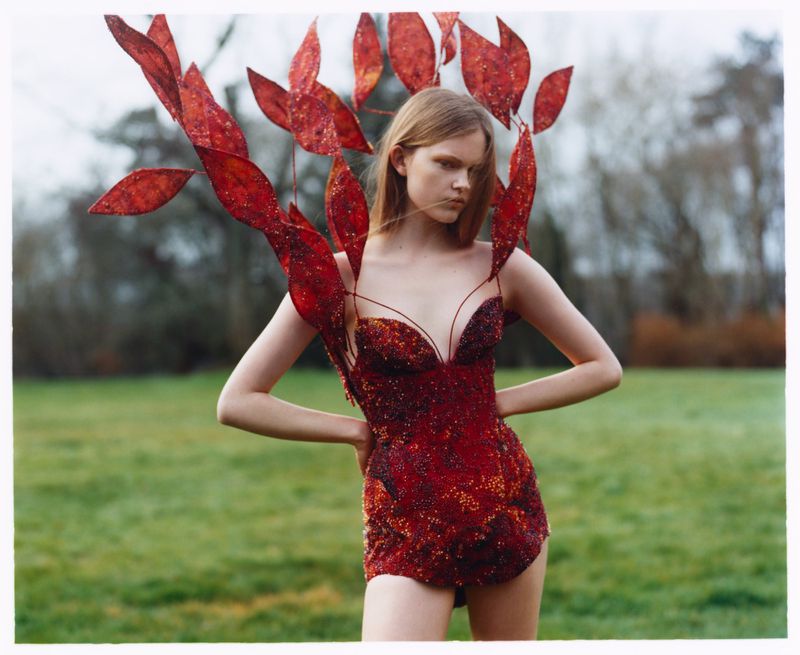 Woman in ornate red dress with leaf-like headdress poses in a grassy field, trees blurred in the background, creating a surreal outdoor fashion image.