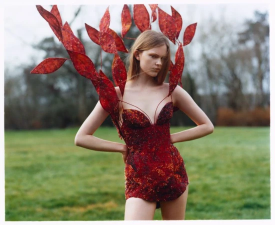Woman in ornate red dress with leaf-like headdress poses in a grassy field, trees blurred in the background, creating a surreal outdoor fashion image.