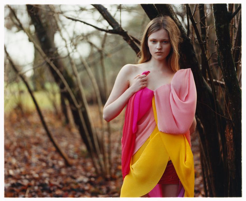 A young woman wearing a multi-coloured pleated garment stands against blurred tree trunks in an outdoor setting.