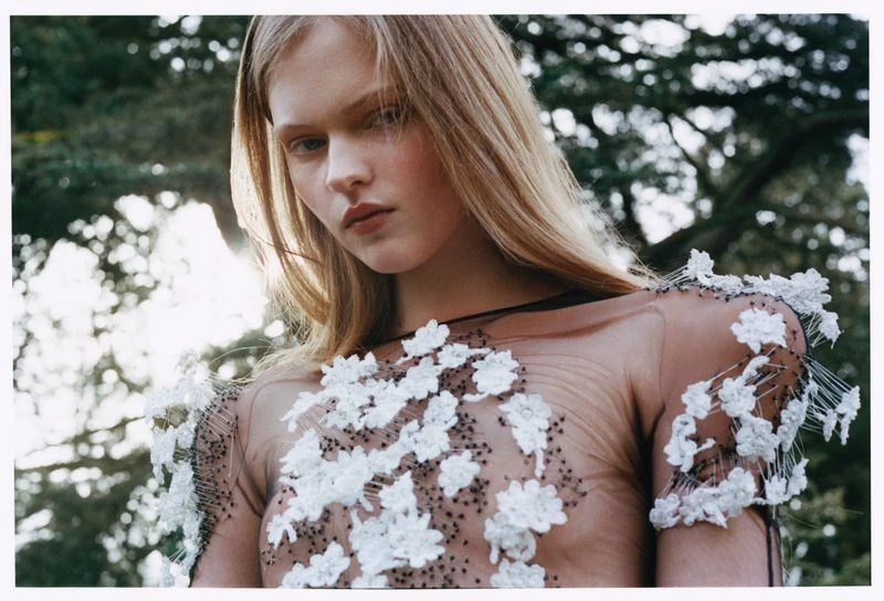 Woman in sheer floral dress posing outdoors. Blonde hair, pale skin, and sunlight filtering through trees create an ethereal mood.