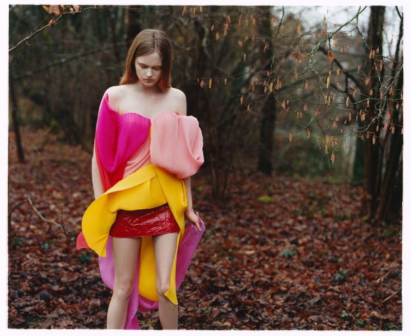 A woman stands in a forest wearing a colorful, avant-garde dress of pink, yellow, and red hues, leaves scattered on the forest floor.