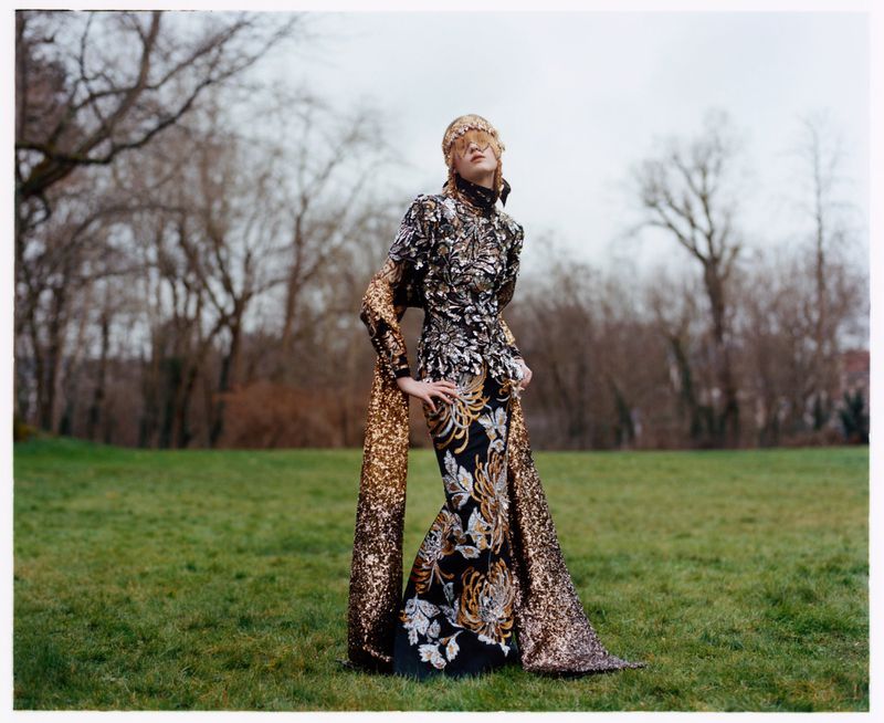 A fashionable woman wearing a long, sequined gown and a headpiece poses outdoors in front of trees in a full-length fashion photograph.