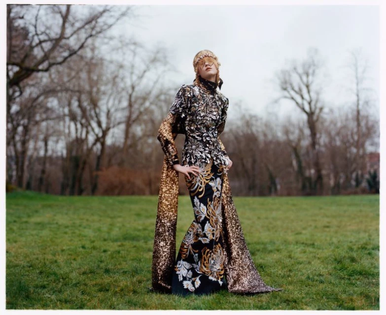 A fashionable woman wearing a long, sequined gown and a headpiece poses outdoors in front of trees in a full-length fashion photograph.