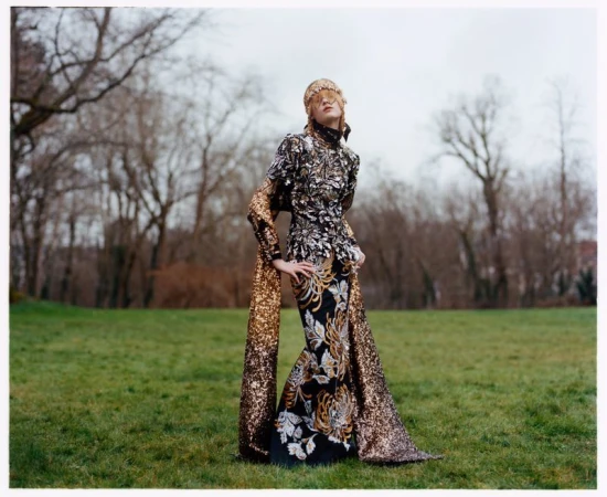 A fashionable woman wearing a long, sequined gown and a headpiece poses outdoors in front of trees in a full-length fashion photograph.