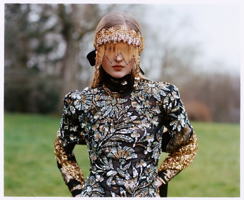 Woman wearing a black and gold embellished dress and beaded headpiece stands in a grassy outdoor setting in this fashion editorial shot.