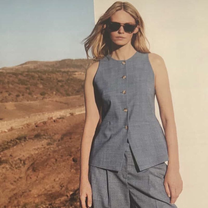 Model wearing dark sunglasses and a light blue sleeveless button-down top against an arid landscape background.